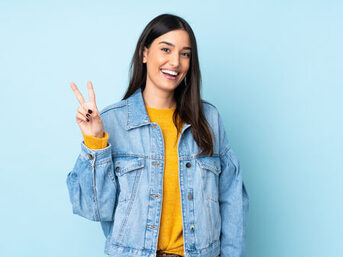 Eine junge Frau steht vor einem blauen Hintergrund und zeigt das Peace-Zeichen in die Kamera. Sie lächelt.