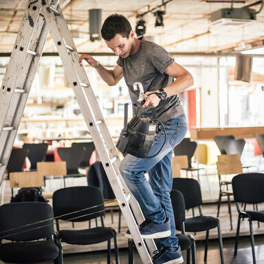 Ein junger Veranstaltungstechniker steht auf einer Leiter und hat einen Scheinwerfer in der Hand.