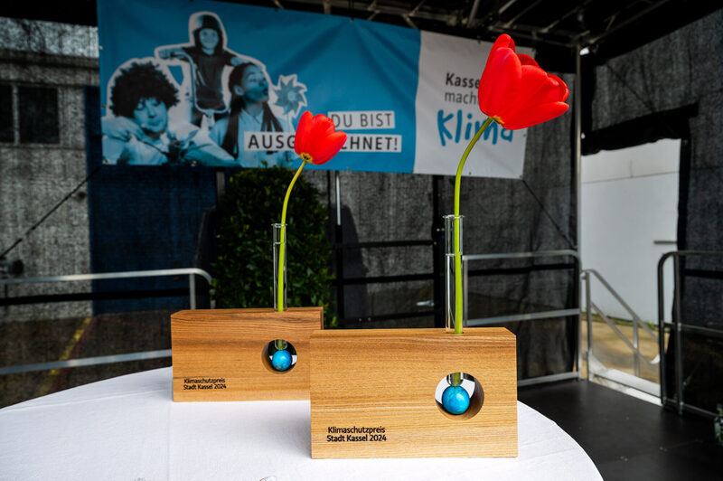 2 Skulpturen aus Holz mit der Schrift "Klimaschutzpreis Stadt Kassel 2024". In der Skulptur befindet sich ein Loch in dem eine Weltkugel liegt, darauf steht ein Reagenzglas in dem eine Tulpe steht.
