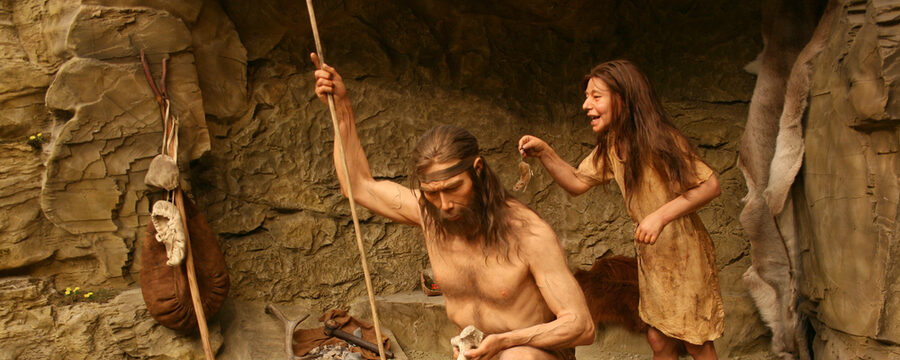 Eine lebensechte Nachbildung eines Mädchens hält eine Maus in den Nacken des vor ihr knienden Vaters in einer Steinzeit-Szene.