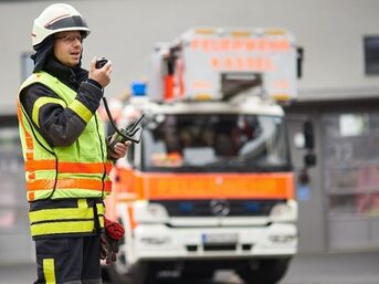 Ein Feuerwehrmann spricht in ein Funkgerät und steht vor einem Feuerwehrauto.
